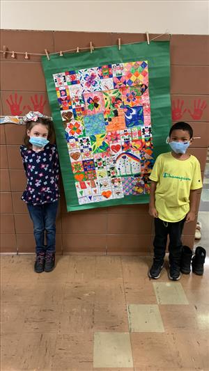 Two students stand on either side of a paper "quilt" hanging from the wall. The quilt is made of squares created by students.